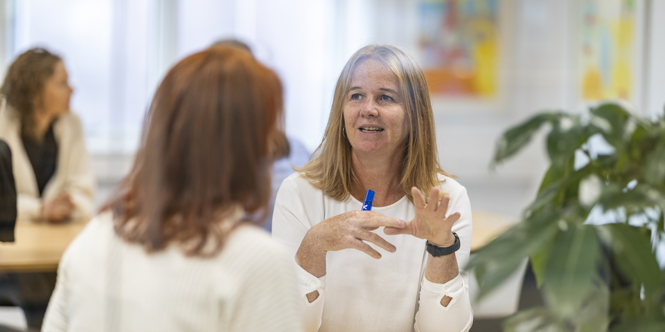 To kvinder sidder og taler sammen ved et bord, hvide trøjer, plante i baggrunden.
