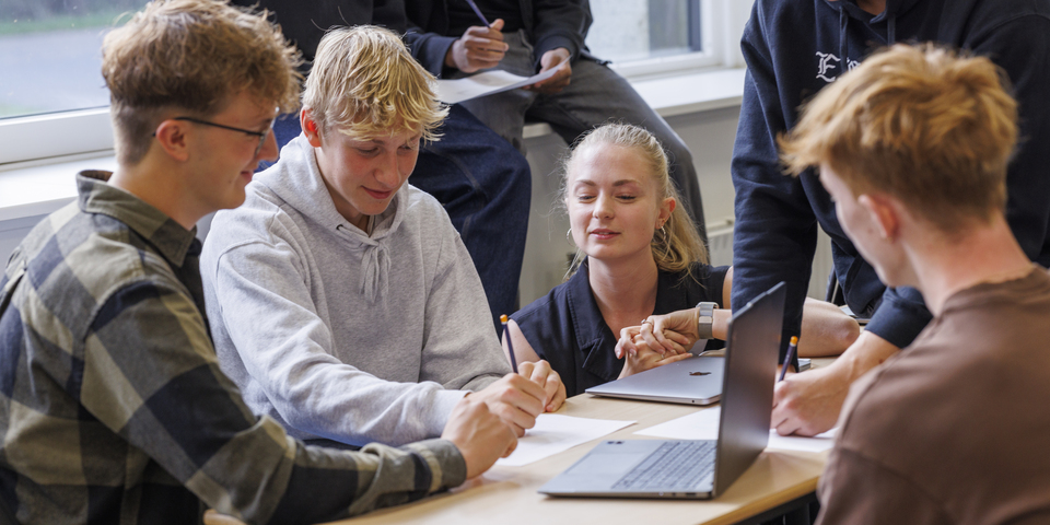 Studerende fra VIA der overtager undervisningen på et gymnasie for en dag. en gruppe af studerende sidder rund om et bord, de skriver noter på et papir og har computere fremme på bordet. To sidder i vindueskarmen med et papir. Smil, samarbejde, studie, noter, undersøgelse, gruppearbejde, ternet skjorte, grå hættetrøje, blyant, macbook, computer, ur, halskæde, briller. 