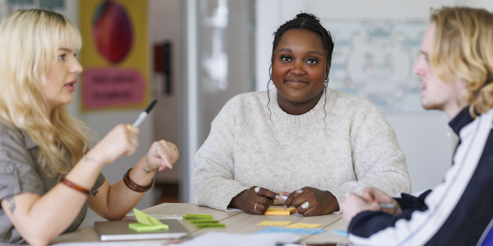 Et gruppearbejde med tre studerende, der noterer på post its. Computer, bærbar, tusser, grå skjorte, halskæde, træbord, armbånd, fingerringe, diversitet, stemning, tusser, træbord, sorte bænke, noter, bøger, læring, blå, hvid, sort sweatshirt. 