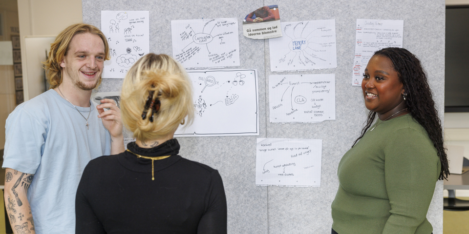 En gruppe på tre studerende står foran en grå opslagstavle, papirer, noter, workshop, brainstorm, samtale, samarbejde, gruppearbejde, hårklemme, halskæde, jeans, udklip, avisudklip, sort stol, bord, grøn langærmet t-shirt, tatoveringer. 