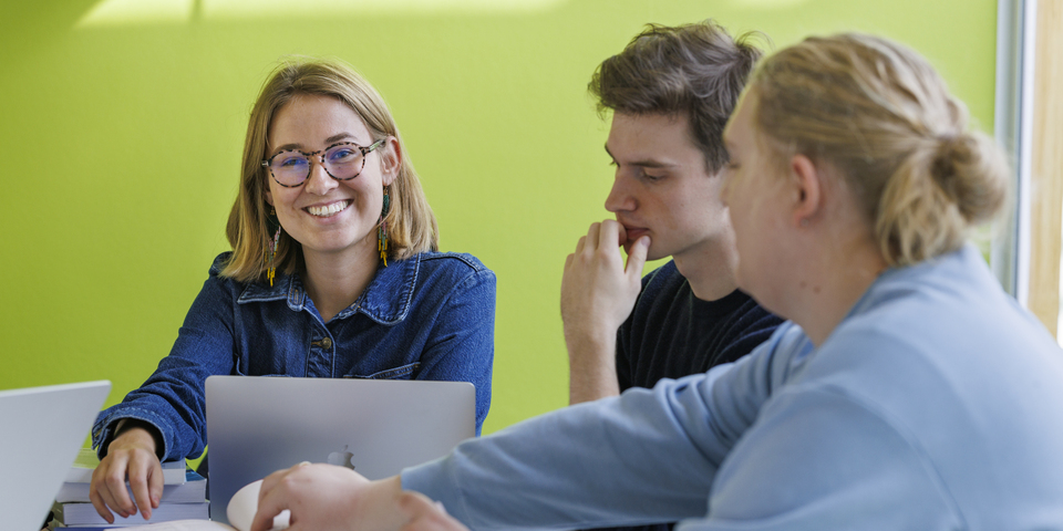 En gruppe af studerende sidder i en lokale med grønne vægge rundt om en computer bærbar, laptop, macbook, på et hvidt bord, hvor der ligger bøger i gul, blå og rød. Denimskjorte, sweatshirt, sneakers, briller, lyst hår, mørkt hår, lysindfald, klistermærke, smil, grin, refleksion, fotograferet gennem en glasvæg. 