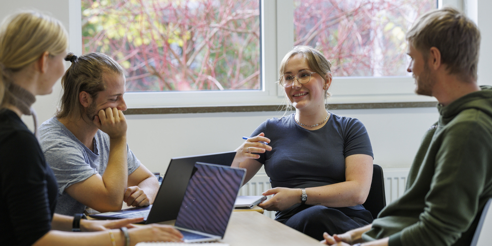 En gruppe af mandlige og kvindelig studerende laver gruppearbejde. Læsning, borde, stole, sorte stole, bærbar, computer, ur, musik i medier, briller, kuglepen, knold, skæg. 