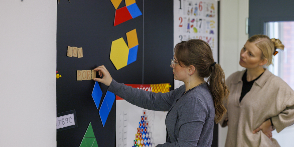 To kvindelige studerende hænger bogstaver på en opslagstavle med farvede figurer mønstre, port, lov, plakat, tal, hestehale, briller, beige sweater, døråbning, grå cardigan, hårklemme. 