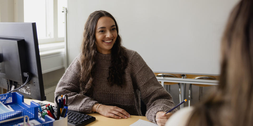 To studerende agerer borger og socialrådgiver i en undervisningssituation. Papir med noter, vinduer, krøllet hår, kontorstole, træbordplade, mørkt hår, brun sweater, kontorstole, saks, tusser i forskellige farver, plante, tastatur, blå brevbakker. 