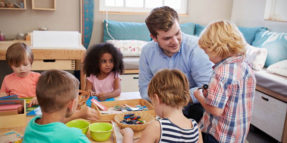 Små børn sidder ved bord  og leger med puslespil og farvekridt sammen med pædagogmedhjælper