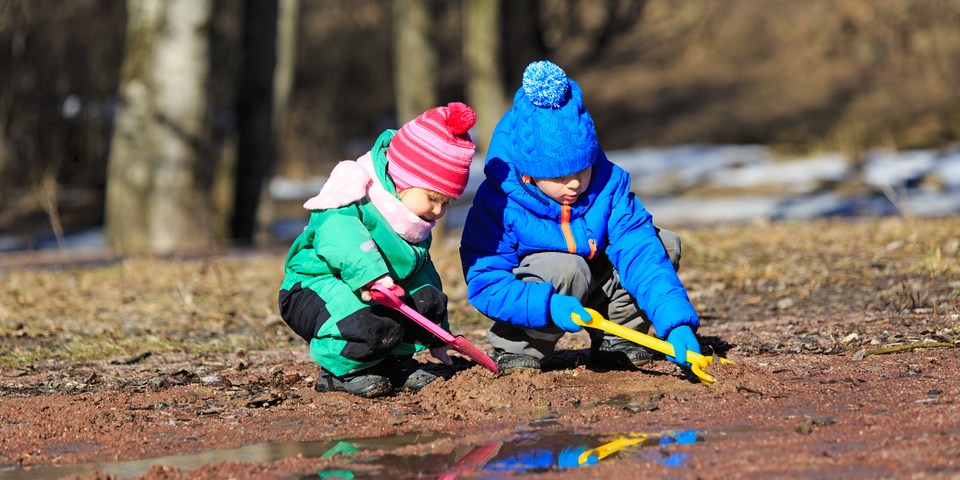 To små børn i flyverdragter leger med skovle ved en vandpyt.
