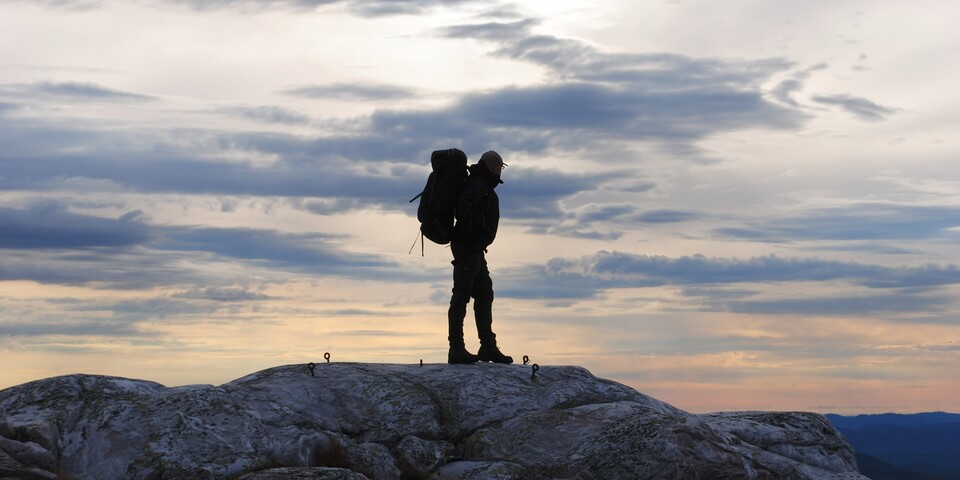 En friluftsvejlederstuderende står med oppakning står på toppen af et fjeld og spejder ud i horisonten. Der er en smuk solnedgang bag personen. 