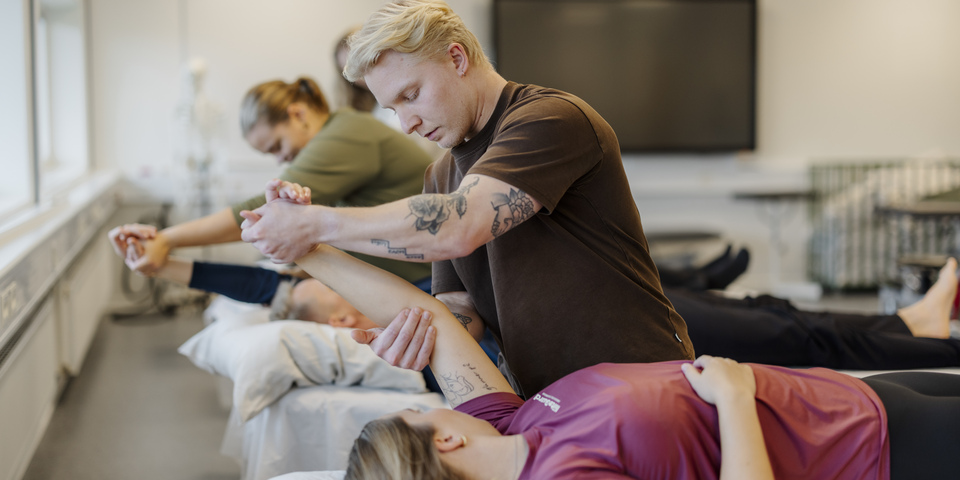 En lokale med fysioterapeutstuderende laver øvelser på hinanden på briks. Skulderøvelser, grå sele, blå elastik, vinduer, skelet, massage, korrektion, briks, lagen, rød t-shirt, tavle, brun t-shirt, tatoveringer, skærm, hovedpude. 