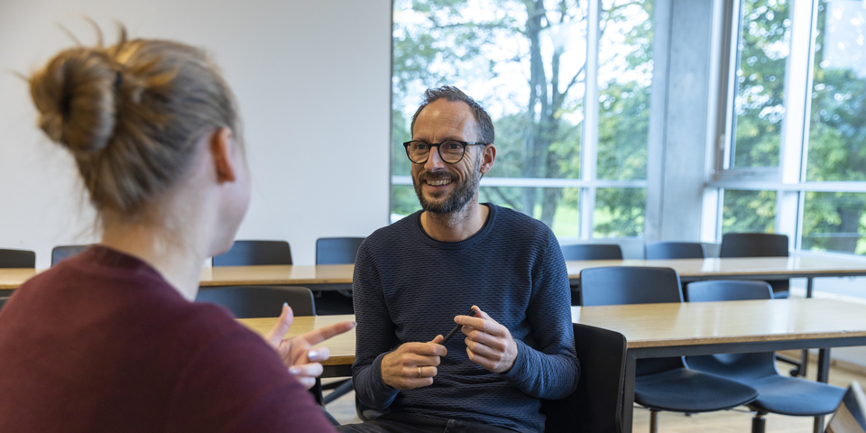 Kvindelig studerende i undervisningslokale får hjælp af lærer / underviser med fagligt indhold. Hun sidder med computer / laptop / pc / bærbar og lytter opmærksomt, nysgerrigt.