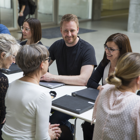 Oppefra / fugleperspektiv af en gruppe smilende og glade personer / kollegaer i gruppearbejde ved bord i atrium på campus. Grønne stole og faglig samtale.