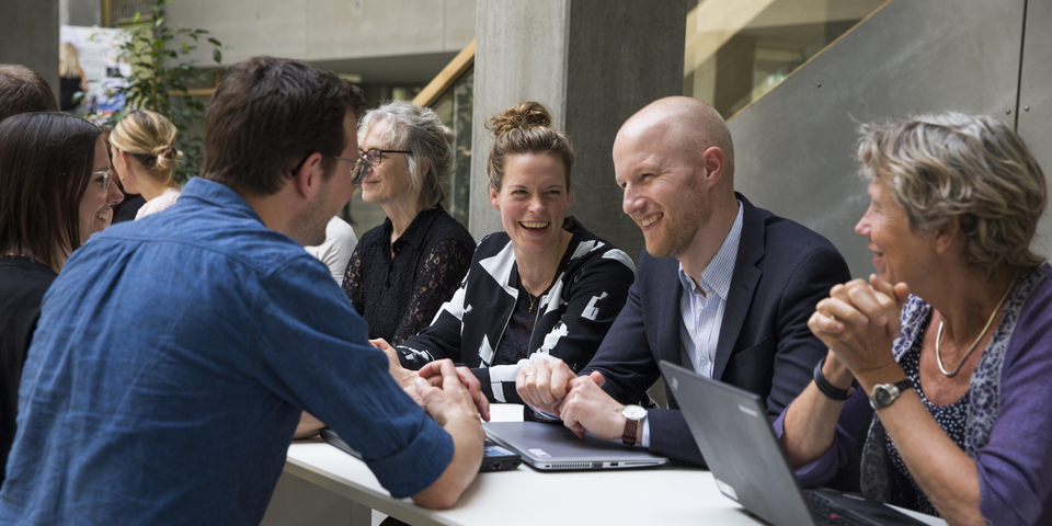En gruppe personer / kollegaer i gruppearbejde til workshop i atrium på campus. Arbejde med computer / laptop ved langbord. Faglig samtale / diskussion. Smil og latter.