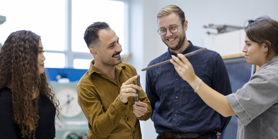 En gruppe af bygningsingeniørstuderende arbejder ved en maskine. Gul skjorte, måling, hænder, knapper, instrumenter, håndtag, observationer, gruppearbejde, metalstang, maskiner, hårklemme, lilla bukser, blå skjorte. 