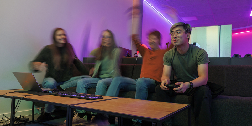 En gruppe af studerende sidder foran en stor skærm med et computerspil i et lokale oplyst af blåt lys. Tilskuerne til spilleren jubler og har hænderne i vejret. Neonfarvede hoveder, VR-briller, gaming, søjle, computer, bærvar, remote, konsol, sneakers, tastatur med blåt lys. 