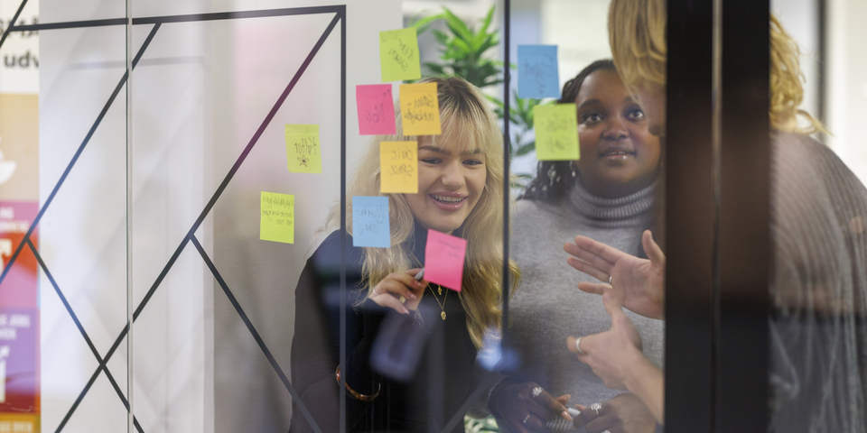 En gruppe af tre studerende laver en øvelse med farvede post-it sedler på en glasvæg, vindue. Jeans, halskæde, gul, blå, pink post its, grønne borde, planter, rødt bordfodboldbord, lang hår, lamper, folie, grafisk design, termokop, diversitet. 