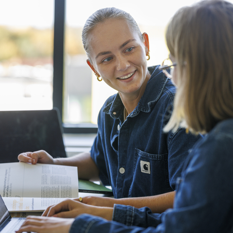 To kvindelige studerende sidder til undervisning og taler sammen mens den ene skriver på en computer, denimskjorte, briller, vindue, sorte stole, bøger, ørering. 