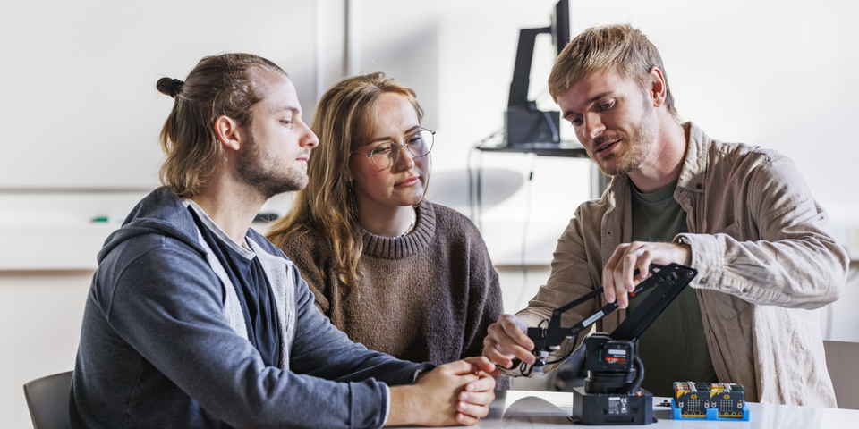 En gruppe af mandlige og kvindelige studerende sidder indenfor med et mikroskop. Tavle, beige stole, hvidt bord, briller, blå sweatshirt, brun sweater, computerskærm. 