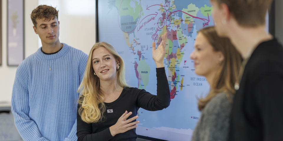To kvindelig studerende og to mandlige studerende peger med en tus foran en skærm i et klasselokale, der viser et verdenskort med forskellige ringe om lande, hvide borde, undervisning, læring, internationalt, præsentation, grå sweater, samtale, blond hår, grå sweater, bærbar, computer. 