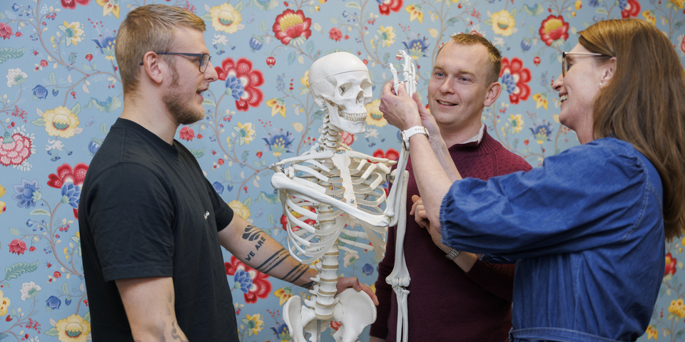 Tre studerende kigger på et skelet, anatomisk model, i undervisningen. Blomstret tapet, denim kjole, sort t-shirt, rød sweater, tatoveringer, dør, briller. Anatomi, knogler, kranie, finger, fingerled, hvidt ur. 