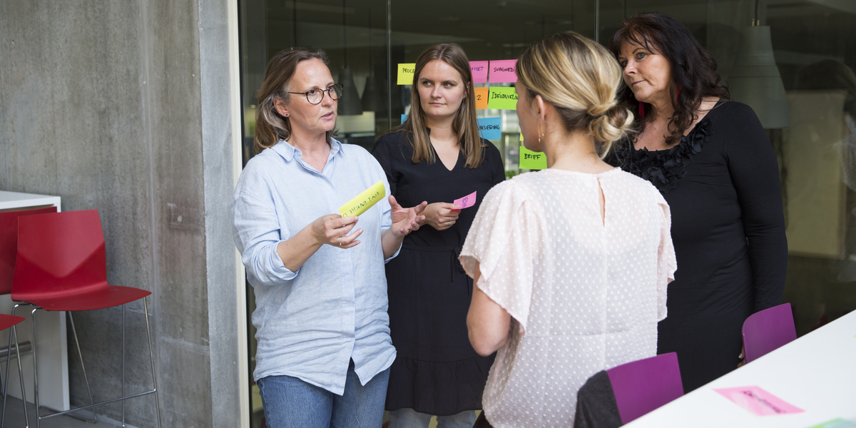 En gruppe af kvinder står med farvede post-it's i hånden, lyserød bluse, briller, rød stol, lilla stol, glasvæg, blå skjorte. 