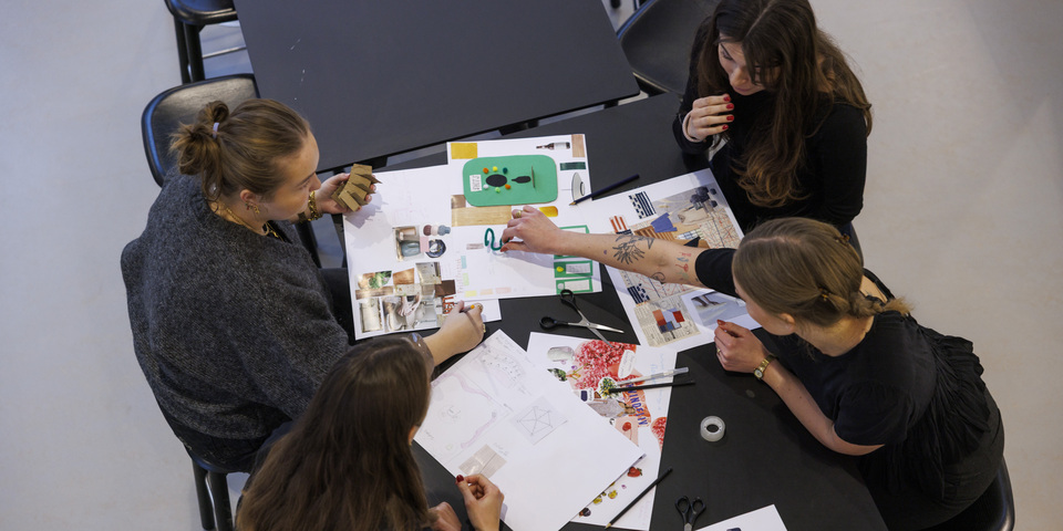 Et gruppearbejde mellem kvindelige studerende foran et træpanel. Bord, moodboards, penalhus, limstift, stole, kuglepen. 