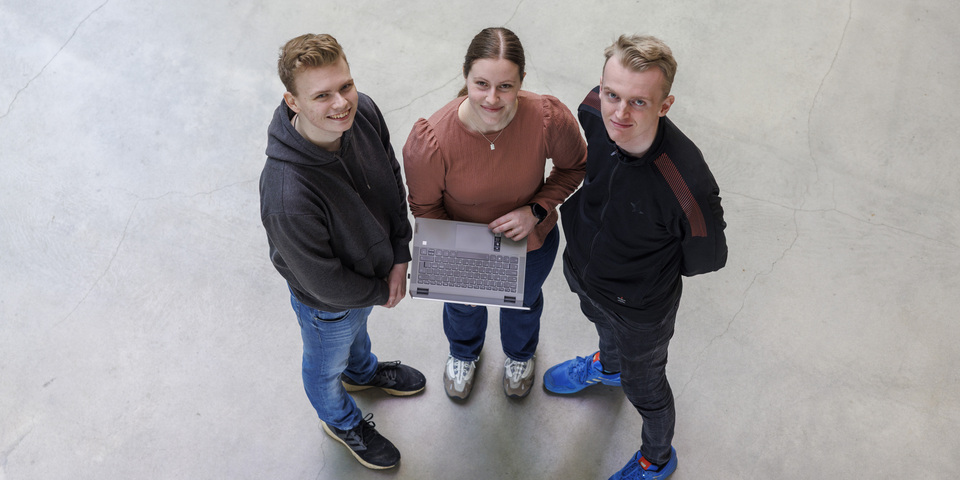 Portræt af tre studerende, der står med en computer i atrium. Rosa bluse, blå sneakers, halskæde, digitalt ur, grå hættetrøje, hvide sneakers. 