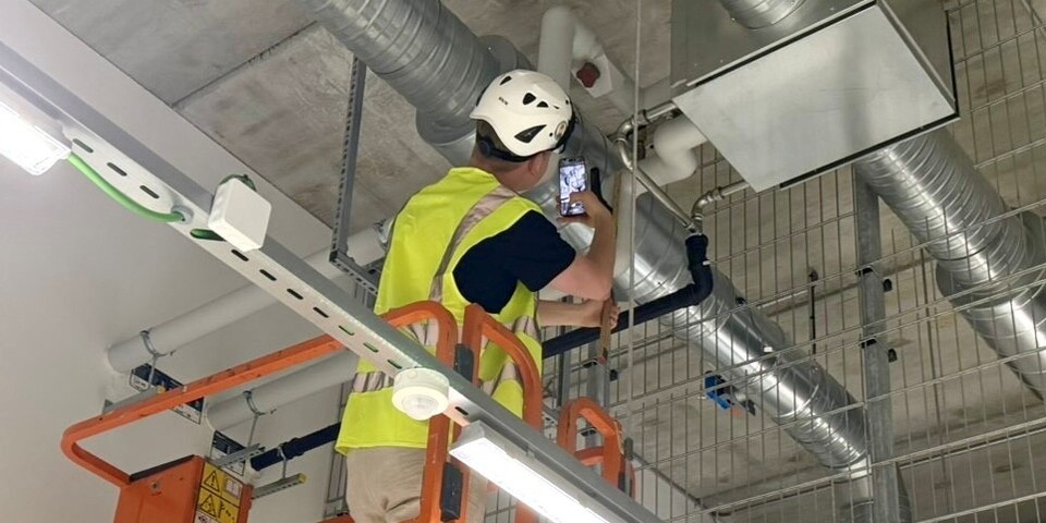 Emil står med ryggen til i en hejst lift med sikkerhedshjelm og gul vest på. Han er ved at kvalitetssikre et ventilationssystem i et rum på en byggeplads.