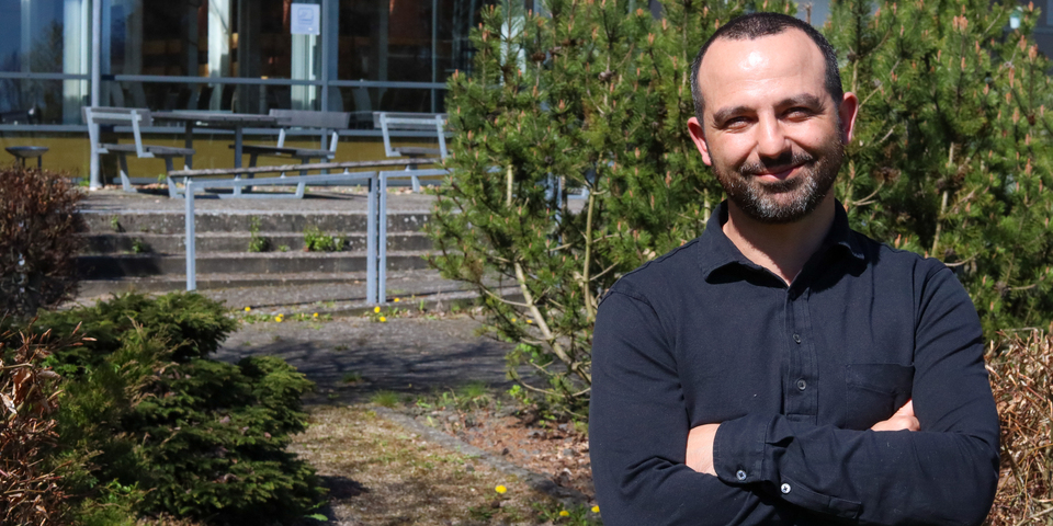 Dennis Can Celik står med armene over kors og smiler ude foran campus Randers. Dennis er iført en sort skjorte og har mørkt skæg og en kortklippet frisure.