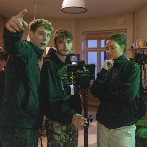 To mandlige og en kvindelig studerende står ved et kamera, fokus, filmoptagelse, sort hættetrøje, samarbejde, gruppearbejde, lamper. 